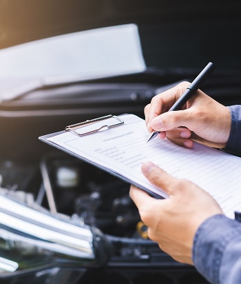 Mechanic writing on a clipboard during vehicle inspection in front of open car hood for collision repair assessment.