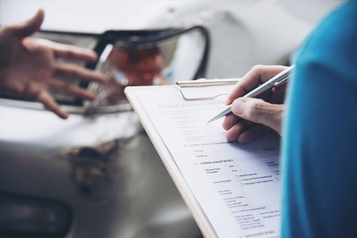 Person filling out auto body insurance claim form after a car accident with visible vehicle damage in background.