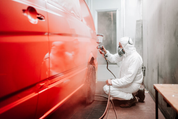Auto technician spray-painting a red car in a professional paint booth.
